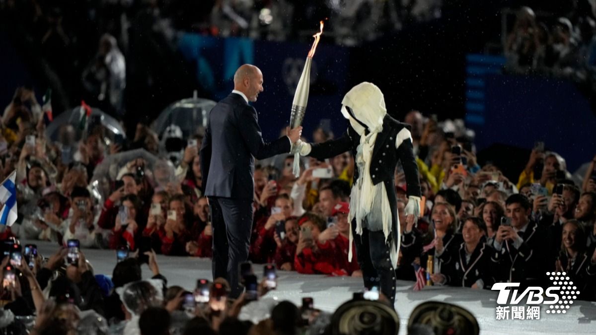 法國傳奇足球明星席丹(Zinedine Zidane)傳遞火炬給一名蒙面人,在典禮現場高舉聖火火炬。 (圖/達志影像美聯社)